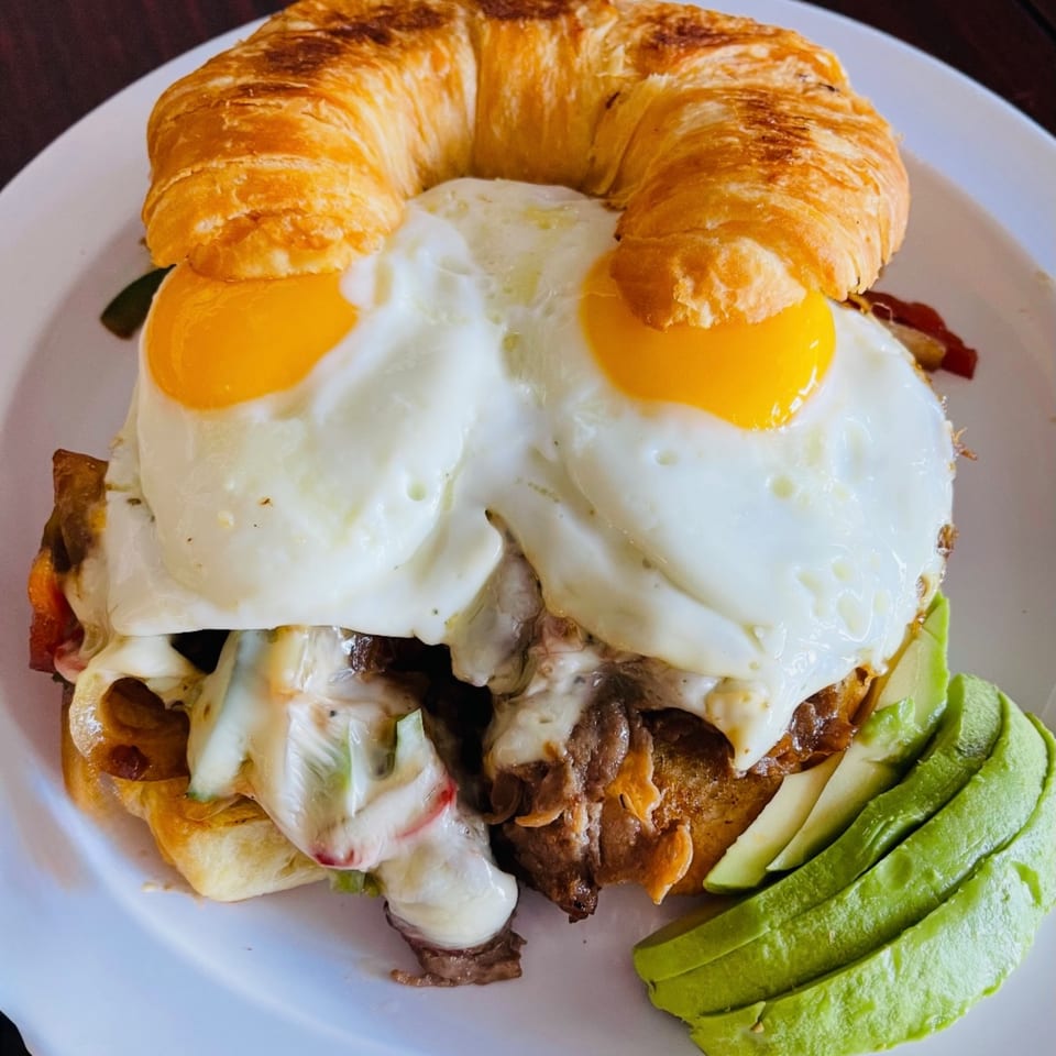 A Sweet Desert Cafe breakfast croissant sandwich with steak, melted cheese, two fried eggs, and avocado.