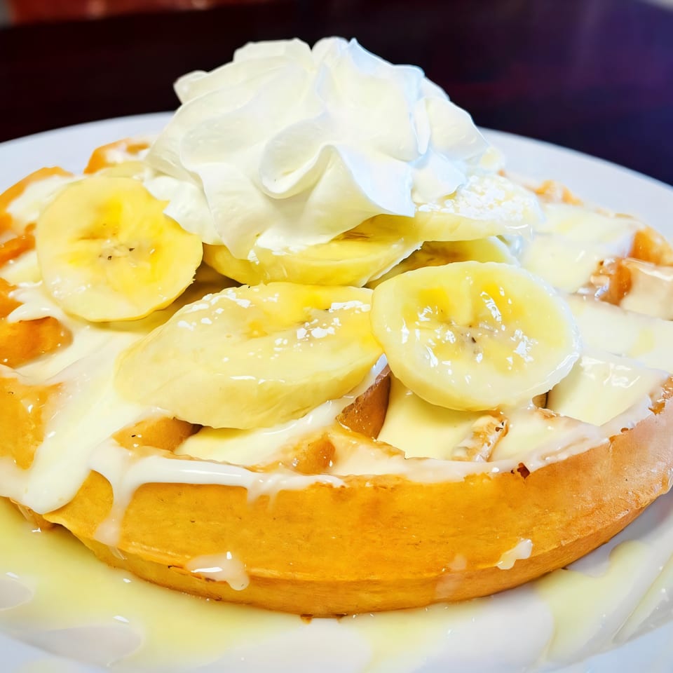 A Sweet Desert Cafe waffle topped with sliced bananas, white sauce, and whipped cream.