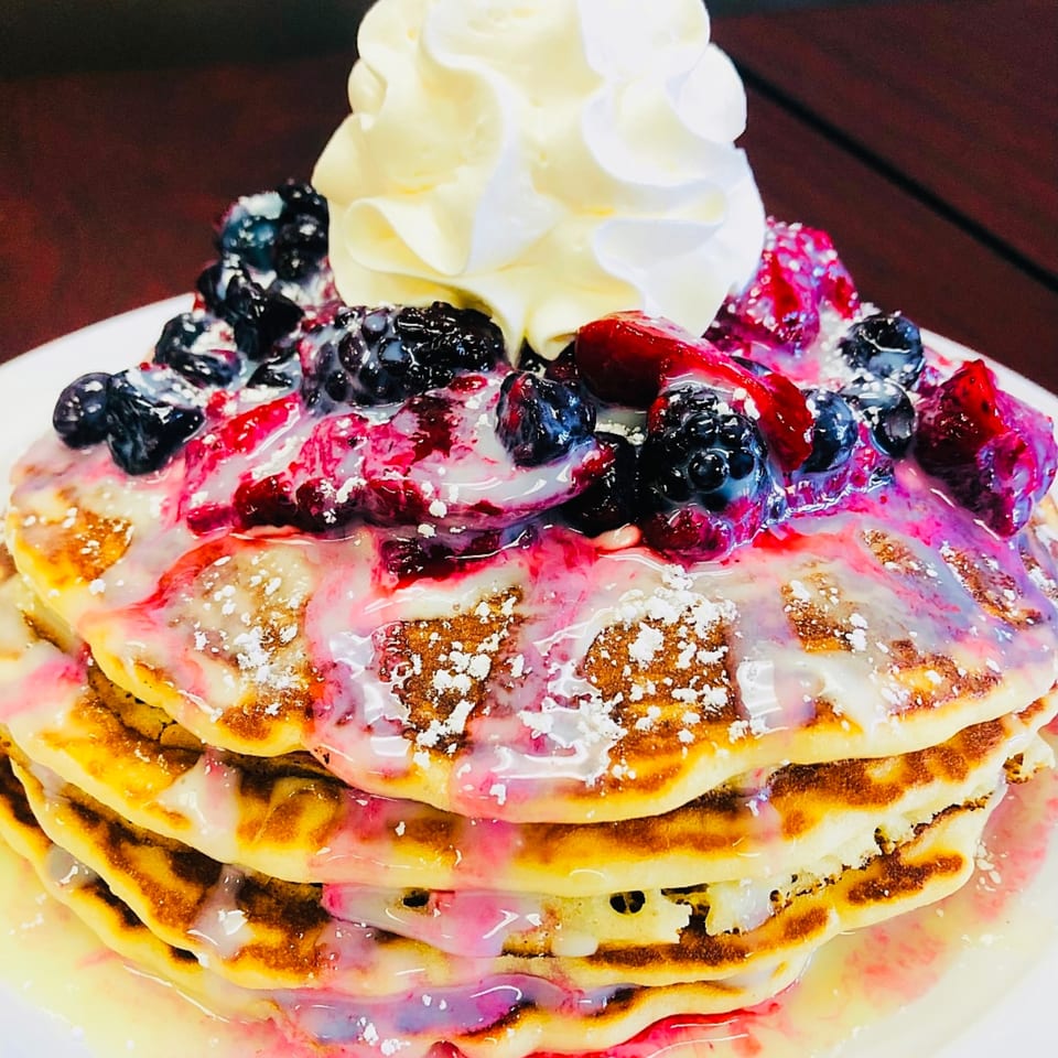 Stack of pancakes topped with mixed berries, white glaze, powdered sugar, and whipped cream.