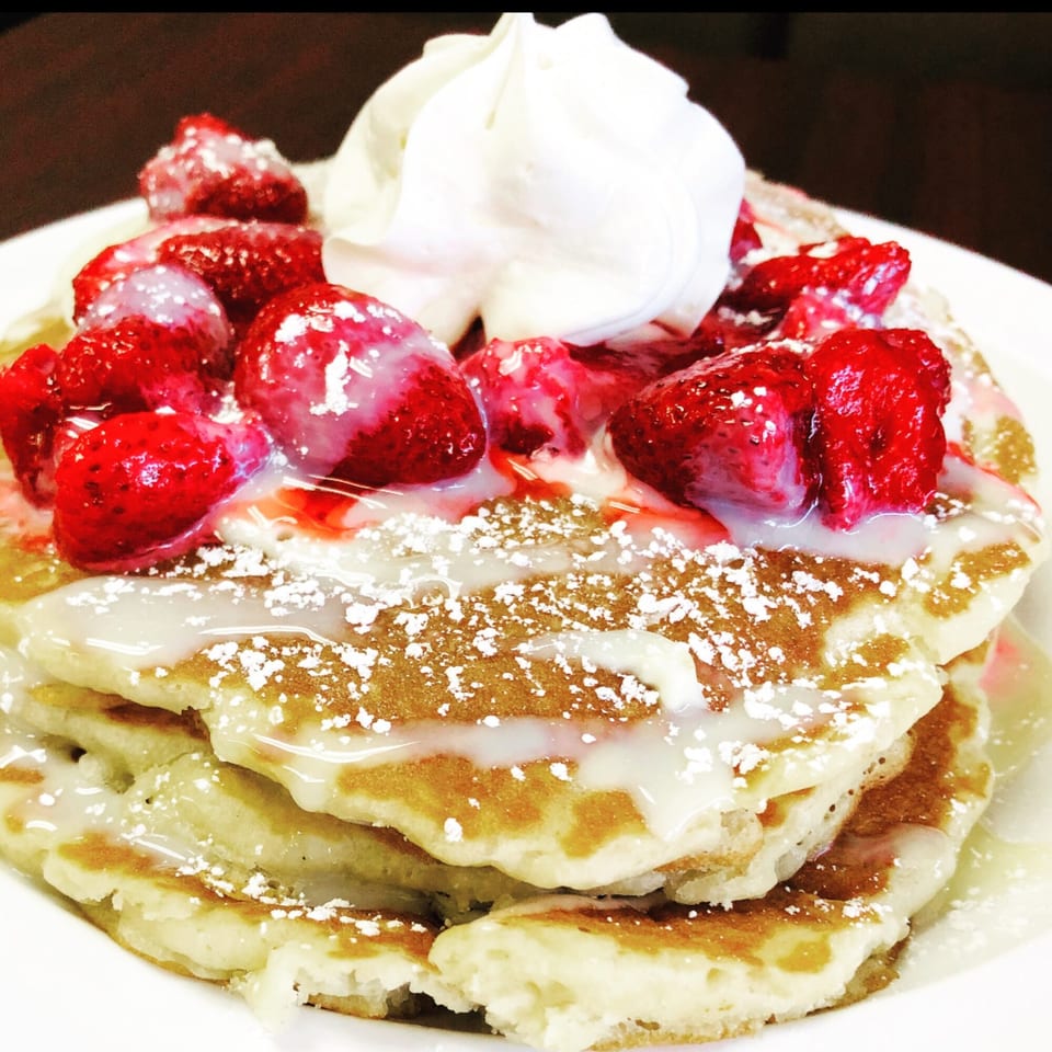 Stack of pancakes topped with fresh strawberries, whipped cream, powdered sugar, and a sweet drizzle.